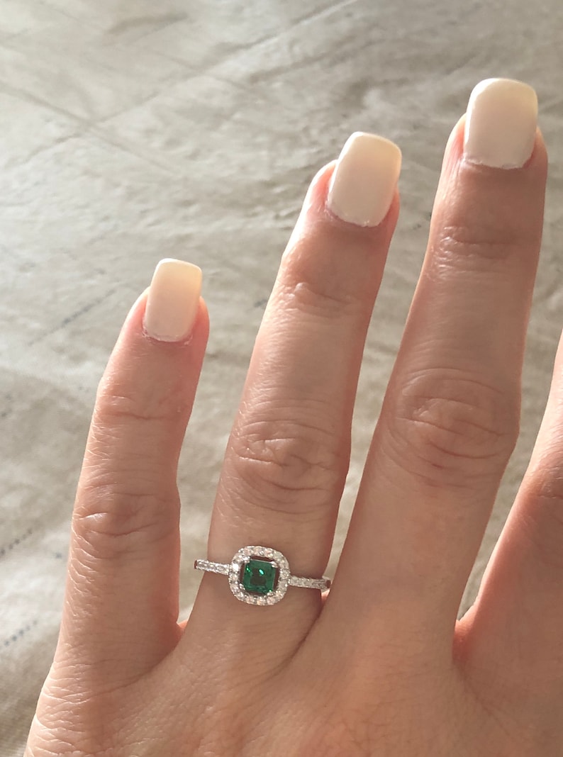 May include: A silver ring with a square emerald green gemstone surrounded by a halo of smaller clear gemstones.