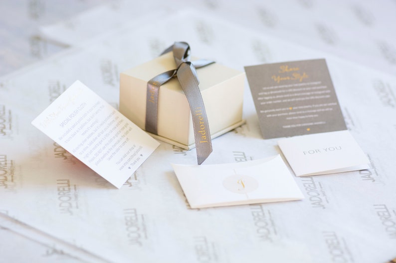 May include: A cream-colored gift box with a gray ribbon and the word "jadorelli" printed on it. Several cards are arranged around the box, including one that says "Show Your Style" and another that says "For You."