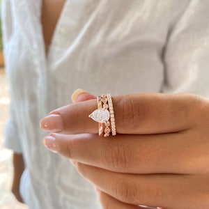 May include: A pear-shaped diamond engagement ring with a rose gold band, set with smaller diamonds. The ring is worn on the finger of a person wearing a white shirt.