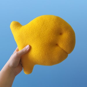 Puede incluir: Un juguete de peluche de pescado amarillo. El pez está hecho de tela suave y peluda y tiene una forma redondeada.