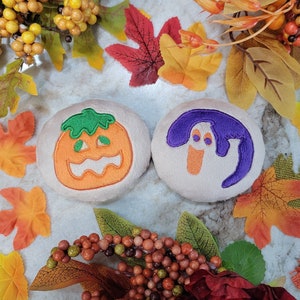 May include: Two plush Halloween-themed toys. One is a pumpkin with a green stem and orange face. The other is a ghost with a purple body and orange eyes.