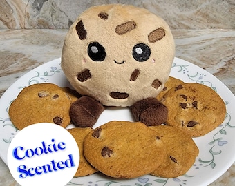 Puede incluir: Un juguete de peluche con forma de galleta con una cara sonriente y detalles de chispas de chocolate, rodeado de galletas de chispas de chocolate reales en un plato blanco con un diseño floral. El plato tiene una pegatina que dice "Cookie Scented".