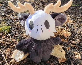 May include: A plush toy in the shape of a deer skull with black fur, large black eyes, and pink cheeks. The toy has large, tan antlers and is sitting on a bed of brown wood chips.
