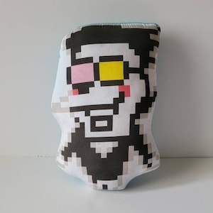 May include: A plush toy shaped like a pixelated character with black hair, a white face, and pink and yellow eyes. The toy is wearing a black shirt and has a blue background.