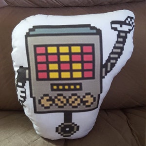 Puede incluir: Una almohada blanca con un diseño de robot pixelado. El robot es gris y tiene una pantalla roja y amarilla. El robot tiene un solo brazo y una sola pierna.