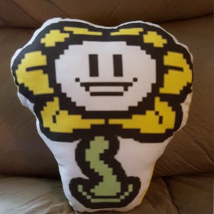 May include: A plush pillow shaped like a smiling yellow flower with a white face and green stem. The flower has a pixelated design, with black eyes and a mouth. The pillow is resting on a brown surface.