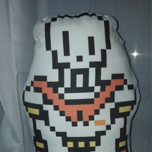 Puede incluir: Un muñeco de peluche blanco con un diseño pixelado de un personaje con una combinación de colores negro, naranja, amarillo y azul. El personaje tiene una cabeza rectangular y una amplia sonrisa.