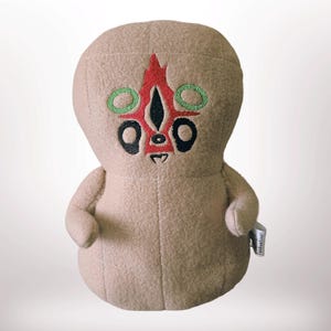 May include: A tan plush toy shaped like a peanut with embroidered facial features. The face includes a red central marking, two green circular eyes, two black oval eyes, and a small black mouth. A small white fabric tag is attached to the side.