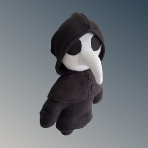 May include: A soft toy plague doctor, with a black hooded cloak and a white beak mask. The doll has two black buttons and is made of fabric. The background is a gradient of grey and white. A unique handmade item.