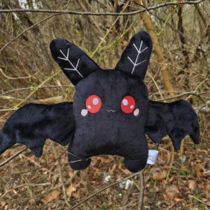 Könnte beinhalten: Ein schwarzes Plüschtier in Form einer Fledermaus mit großen roten Augen, kleinen rosa Wangen und weißen blattartigen Designs auf den Ohren. Die Fledermaus hat ausgebreitete Flügel.