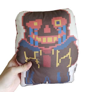 May include: A rectangular plush pillow with a pixelated character design. The design features a brown body, red and yellow accents, and blue details. The pillow is held against a white background.
