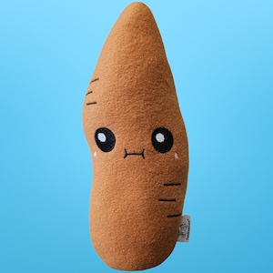 May include: A brown plush toy shaped like a sweet potato, with a cartoon face featuring large black eyes, a small mouth, and pink cheeks. The toy has three horizontal lines on the side and is set against a light blue background.