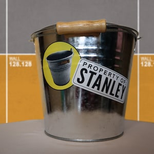 Può includere: Un secchio di metallo argentato con un manico di legno. Il secchio ha un adesivo che dice "Proprietà di: Stanley" e un adesivo più piccolo di un secchio argentato.