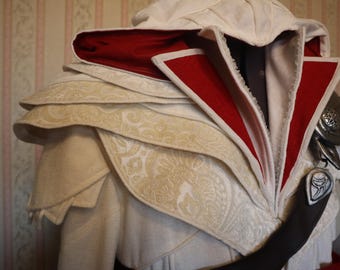May include: A white hooded cloak with a red lining and intricate white embroidery. The cloak has a layered design and features a decorative metal piece on the shoulder.