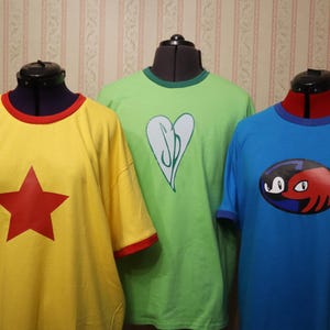 Op de afbeelding: Drie t-shirts op mannequins. Een geel shirt met een rode ster, een groen shirt met een wit hartontwerp en een blauw shirt met een rood-zwarte afbeelding. Elk shirt heeft contrasterende randen.