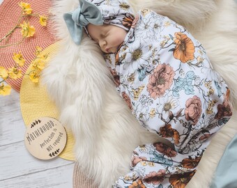 May include: A baby swaddled in a white floral swaddle with a light blue and white knotted headband. The baby is sleeping on a white fluffy surface. A wooden sign with the text "Motherhood. Perfection is not required." is in the foreground.