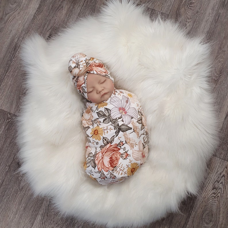 May include: A newborn baby swaddled in a floral print swaddle with a matching floral headband. The swaddle is a light pink, yellow, and green with a floral pattern. The headband is a light pink with a floral pattern.