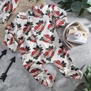 Peut inclure: Une grenouillère blanche pour bébé avec un motif de cœur rouge et de feuilles vertes. La grenouillère porte le texte "Mama's Boy" répété sur tout le design.