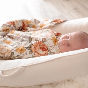 May include: A newborn baby sleeping peacefully in a white, cushioned nest with a floral patterned swaddle blanket.
