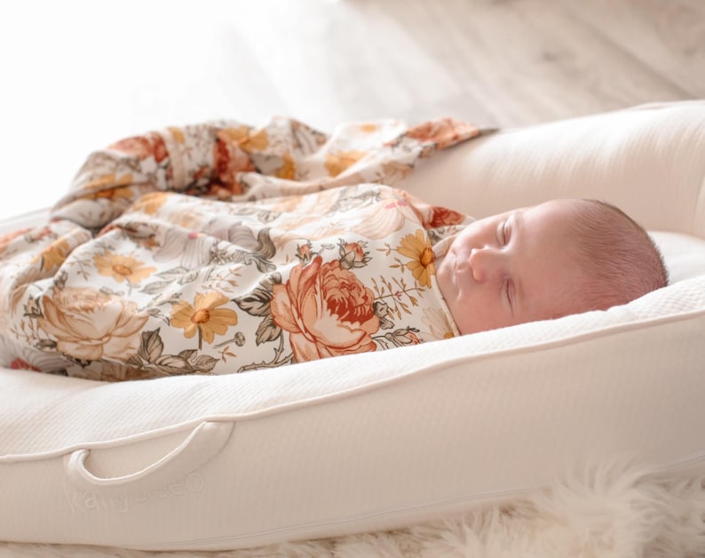 May include: A newborn baby sleeping peacefully in a white, cushioned nest with a floral patterned swaddle blanket.