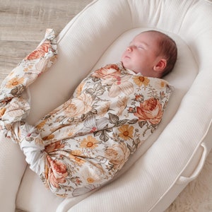 May include: A baby swaddled in a white and floral patterned swaddle, sleeping in a white, oval-shaped, padded baby nest.