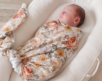May include: A baby swaddled in a white and brown floral swaddle, sleeping in a white, oval-shaped, padded bassinet.