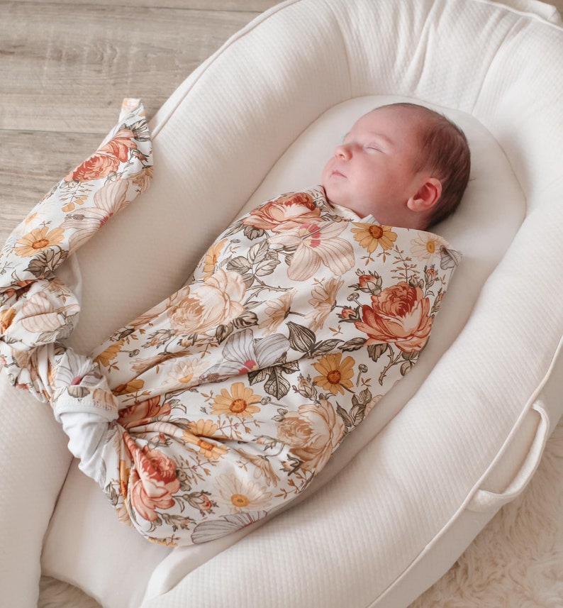 May include: A baby swaddled in a white and floral patterned swaddle, sleeping in a white, oval-shaped, padded baby nest.