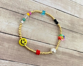 May include: A gold beaded bracelet with colorful charms, including a smiley face, a flower, and a pearl.