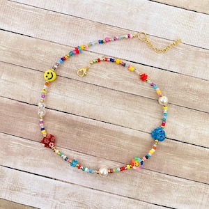 May include: A colorful beaded necklace with a gold chain clasp. The necklace features various charms, including a smiley face, a flower, a heart, and a blue star. The necklace is made with small, colorful beads and a single white pearl.