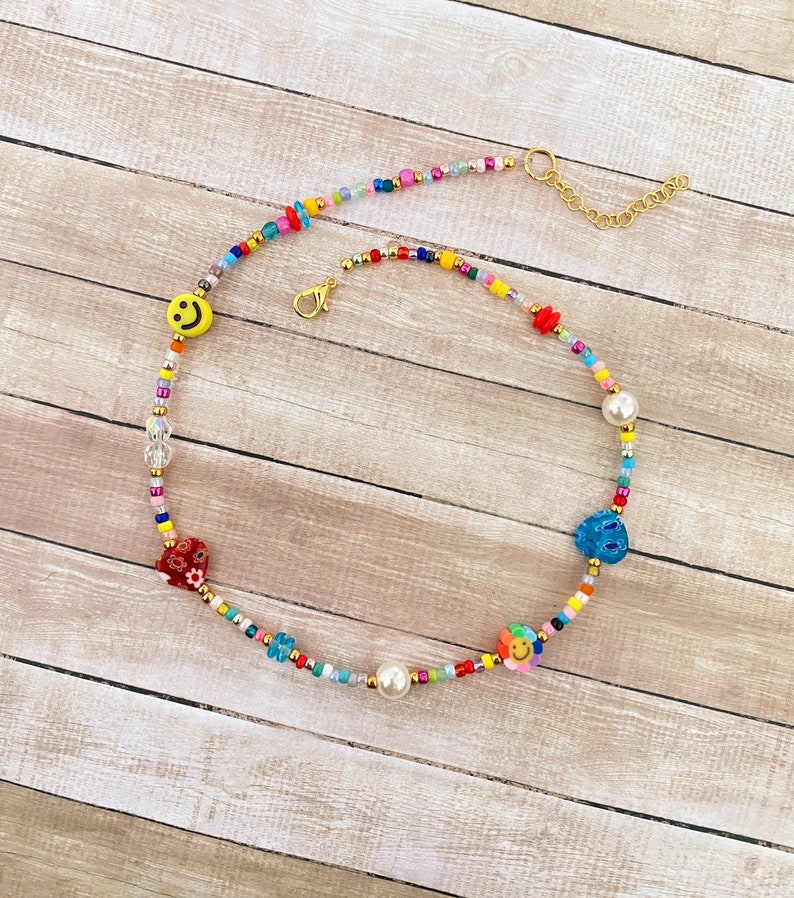 May include: A colorful beaded necklace with a gold chain clasp. The necklace features various charms, including a smiley face, a flower, a heart, and a blue star. The necklace is made with small, colorful beads and a single white pearl.