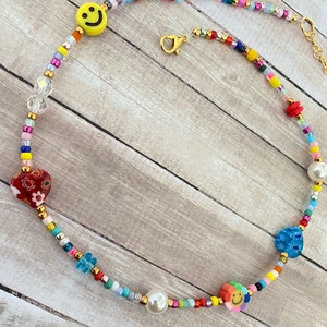 May include: A colorful beaded necklace with a smiley face charm, a red heart charm, a blue flower charm, and a pearl bead. The necklace is made with a variety of small beads in different colors, including red, yellow, blue, green, and white.
