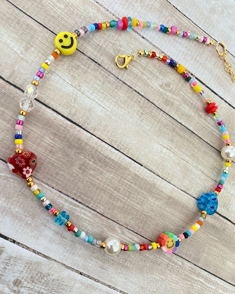 May include: A colorful beaded necklace with a smiley face charm, a red heart charm, a blue flower charm, and a pearl bead. The necklace is made with a variety of small beads in different colors, including red, yellow, blue, green, and white.