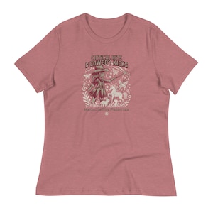 May include: Dusty rose t-shirt with a graphic featuring a cowgirl with a wand, a unicorn, and the text "CRITICAL HITS & COWBOY KICKS MAGIC IN THE FRONTIER". The design is in shades of beige and brown.