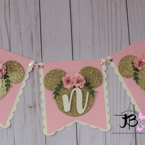 Puede incluir: Bandera de brillo rosa y dorado con las letras "ONE" en brillo dorado, cada letra est&aacute; rodeada de una cabeza de Mickey Mouse de brillo dorado con flores rosas y hojas verdes.