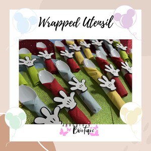 May include: Wrapped utensil sets featuring white spoons and colorful napkins in red, yellow, green, and blue. Each set is adorned with a Mickey Mouse hand-shaped napkin ring. The text "Wrapped Utensil" is at the top.