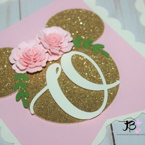 Puede incluir: Bandera de brillo rosa y dorado con una letra blanca "O" y dos flores de papel rosa. La bandera est&aacute; decorada con hojas verdes.