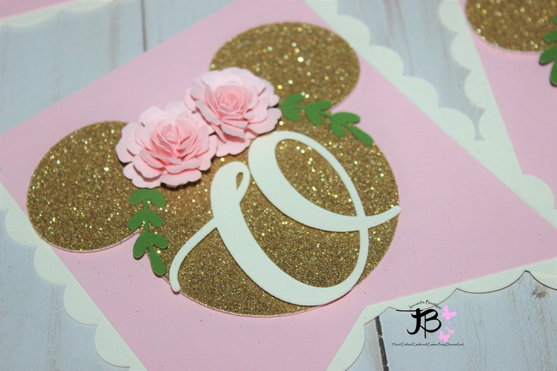 Puede incluir: Bandera de brillo rosa y dorado con una letra blanca "O" y dos flores de papel rosa. La bandera est&aacute; decorada con hojas verdes.