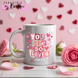 Puede incluir: Una taza de cerámica blanca con las palabras "YOU ARE SO LOVED" en un estilo collage con corazones y estrellas rosas. El texto "JOHN 3:16" está debajo. La taza tiene un asa gris y está rodeada de rosas rosas y confeti en forma de corazón.