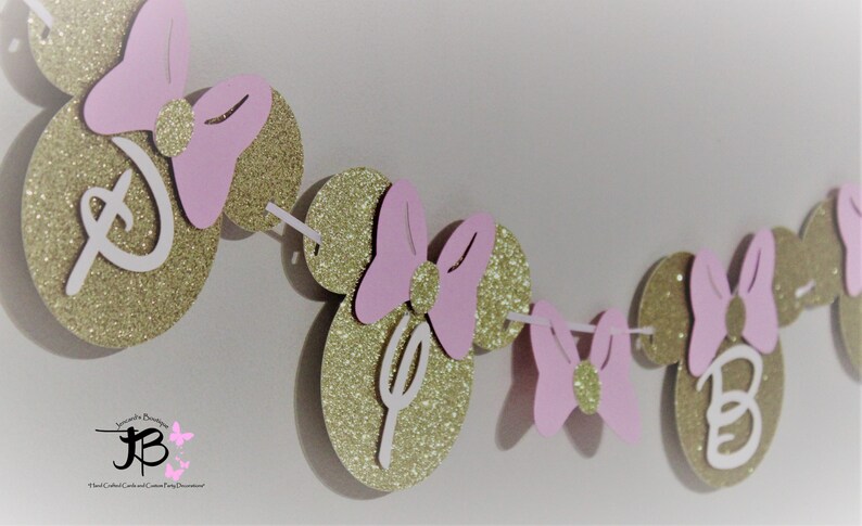 May include: Gold glitter Minnie Mouse ears with pink bows and white letters spelling "D-I-S-N-E-Y" for a birthday party decoration.