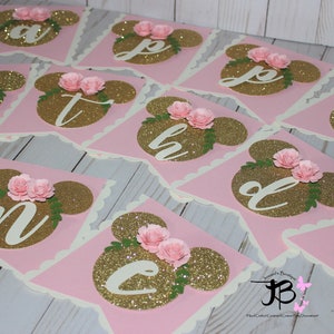 Puede incluir: Bandera de brillo rosa y dorado con orejas de Mickey Mouse y flores. Cada bandera tiene una letra del alfabeto en brillo dorado. La pancarta dice "Feliz cumplea&ntilde;os".