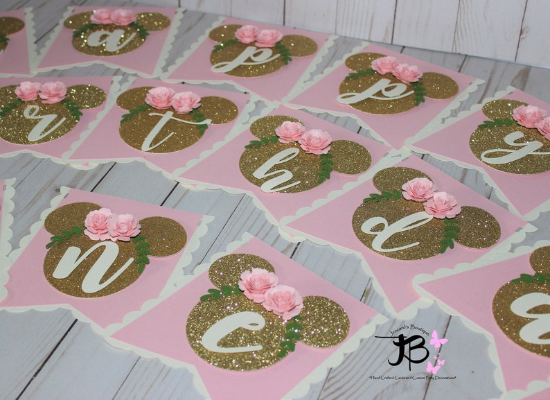 Puede incluir: Bandera de brillo rosa y dorado con orejas de Mickey Mouse y flores. Cada bandera tiene una letra del alfabeto en brillo dorado. La pancarta dice "Feliz cumplea&ntilde;os".