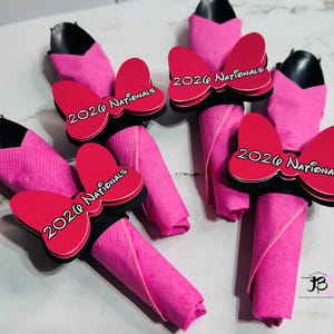 Op de afbeelding: Verschillende roze servetten zijn gerangschikt met zwarte houders en rode strikvormige decoraties. Elke strik heeft de tekst "2026 Nationals" in het wit. De servetten zijn opgerold en staan rechtop, wat een feestelijke presentatie cre&euml;ert.