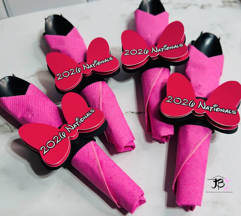 Op de afbeelding: Verschillende roze servetten zijn gerangschikt met zwarte houders en rode strikvormige decoraties. Elke strik heeft de tekst "2026 Nationals" in het wit. De servetten zijn opgerold en staan rechtop, wat een feestelijke presentatie cre&euml;ert.