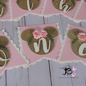 Puede incluir: Bandera de brillo rosa y dorado con letras blancas que deletrean "One" con un dise&ntilde;o de cabeza de Mickey Mouse y flores rosas en cada letra.