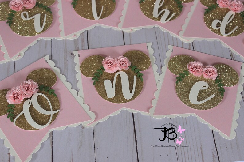 Puede incluir: Bandera de brillo rosa y dorado con letras blancas que deletrean "One" con un dise&ntilde;o de cabeza de Mickey Mouse y flores rosas en cada letra.