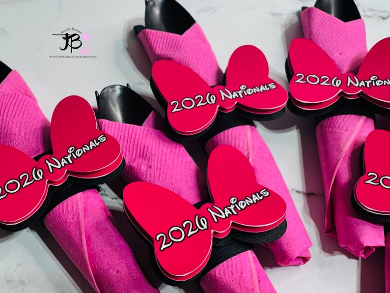 Op de afbeelding: Roze servetringen met een rood vlinderdasontwerp. Elke strik heeft de tekst "2026 Nationals" in het wit. De servetten zijn gevouwen en bevatten zwart keukengerei.