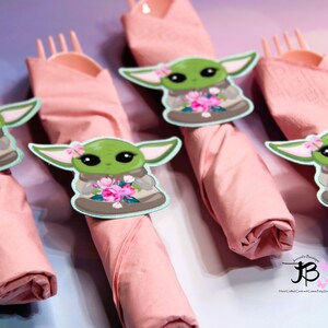 Puede incluir: Cuatro servilletas rosas con un personaje verde que sostiene flores en cada una. El personaje es una versi&oacute;n de dibujos animados de Baby Yoda de la serie Star Wars. Las servilletas est&aacute;n enrolladas y atadas con un tenedor de pl&aacute;stico.