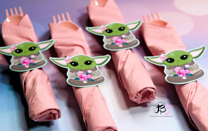Puede incluir: Cuatro servilletas rosas con un personaje verde que sostiene flores en cada una. El personaje es una versi&oacute;n de dibujos animados de Baby Yoda de la serie Star Wars. Las servilletas est&aacute;n enrolladas y atadas con un tenedor de pl&aacute;stico.