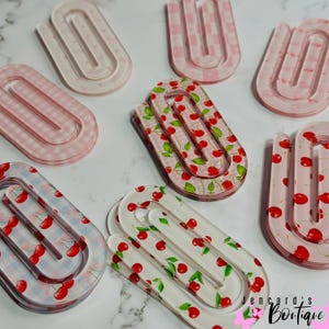 Op de afbeelding: Een verzameling extra grote papieren clips van acryl met diverse patronen, waaronder roze gingham, lichtblauw gingham en rode kersenprints op transparante en witte achtergronden, in een verspreide opstelling.
