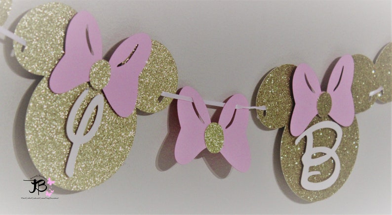 May include: A pink and gold glitter Minnie Mouse themed banner with the letter "B" in the center. The banner is strung together with white string.